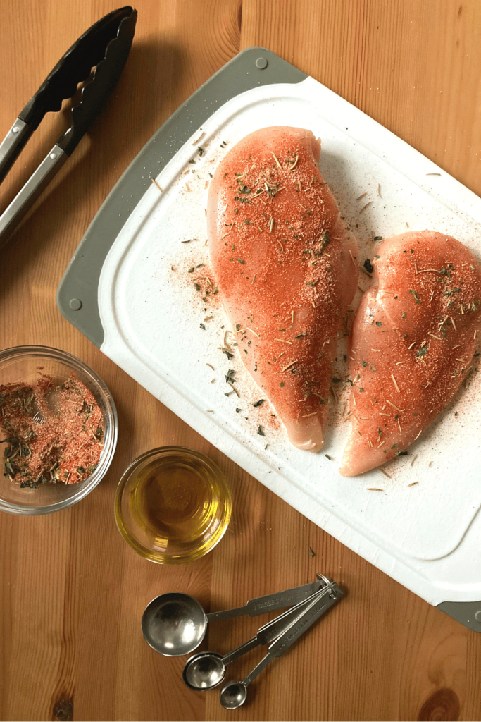 2 chicken breasts on a white cutting board, sprinkled with paprika, garlic powder and rosemary