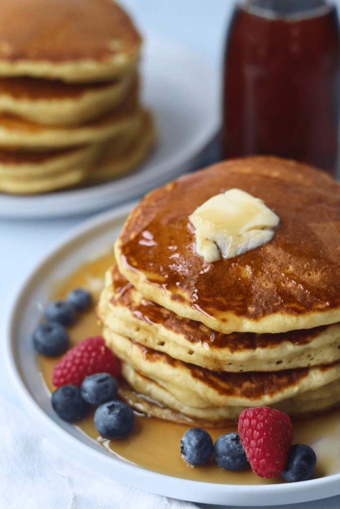 nicely stacked of pancake on a white plate with berries on the side and butter on top and sprinkled maple syrup